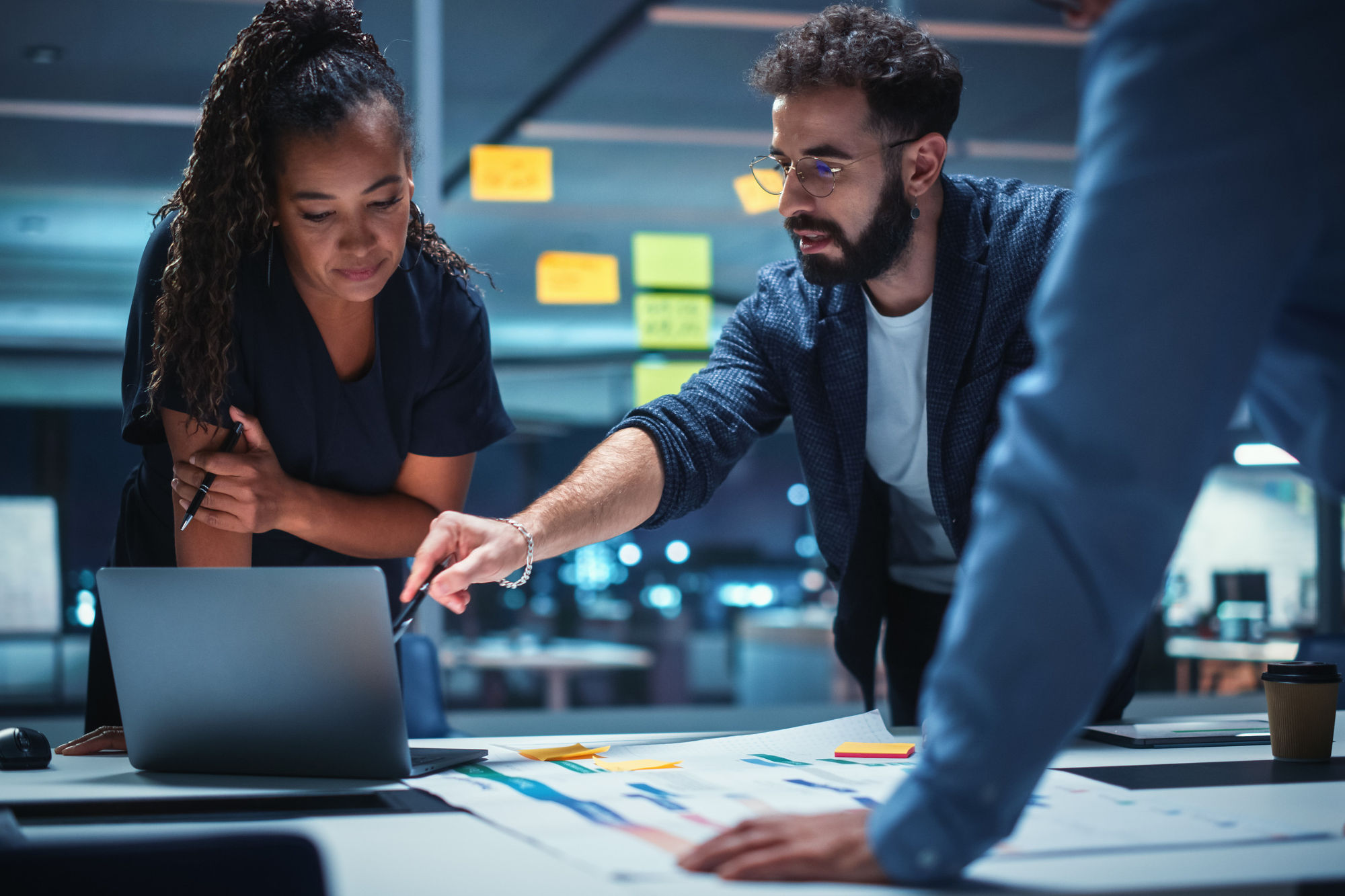A diverse team collaborates around a laptop in a dimly lit office, focused and engaged. 